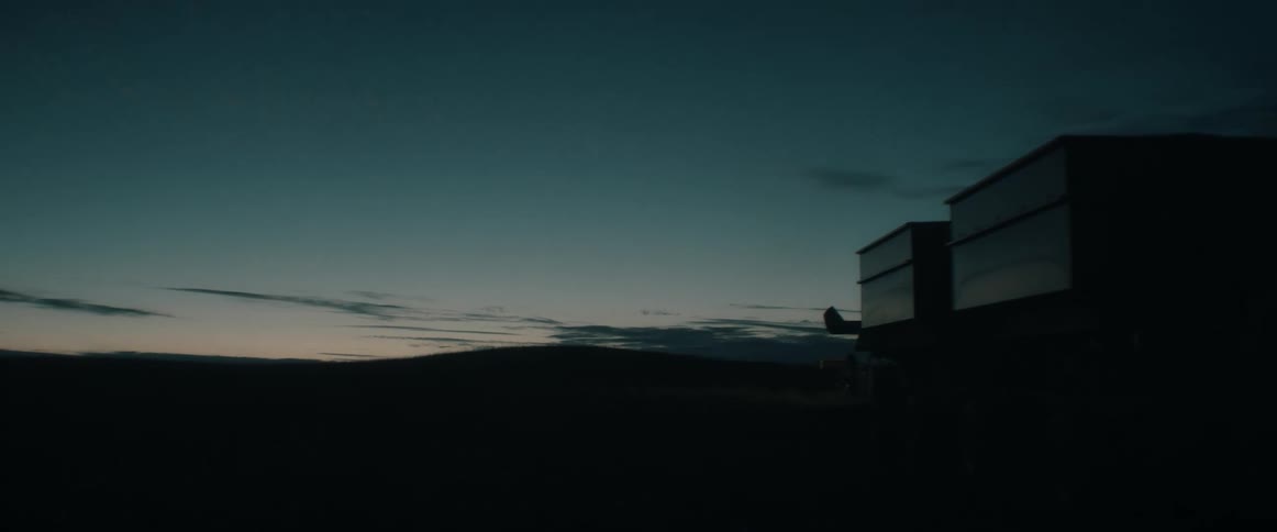 A combine harvester drives away in a dark field with its lights on at twilight