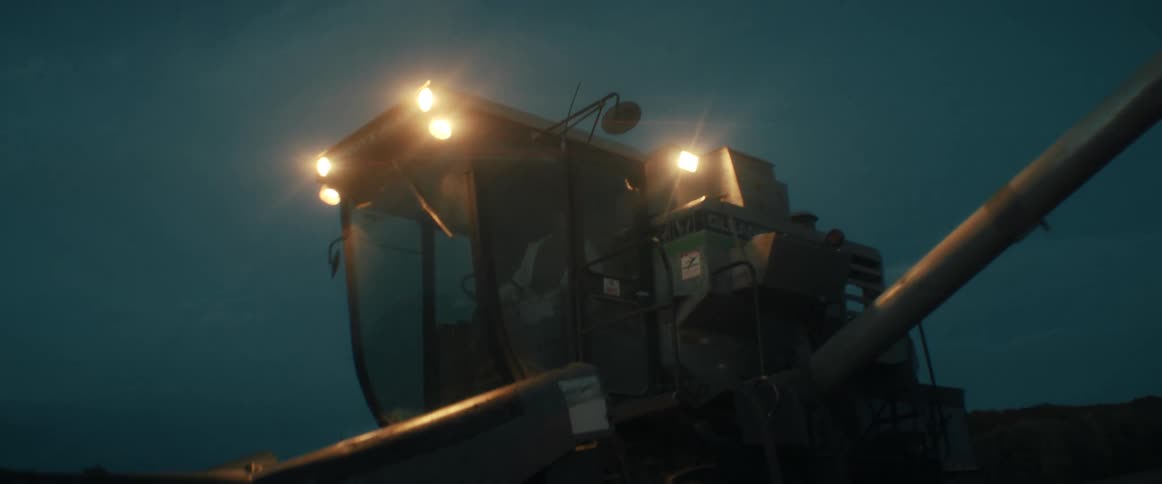 A slow zoom out reveals a large combine harvester with its lights on, working in a field after sunset