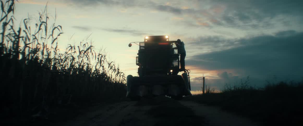 A silhouetted farmer climbs onto his combine harvester next to a cornfield at dusk