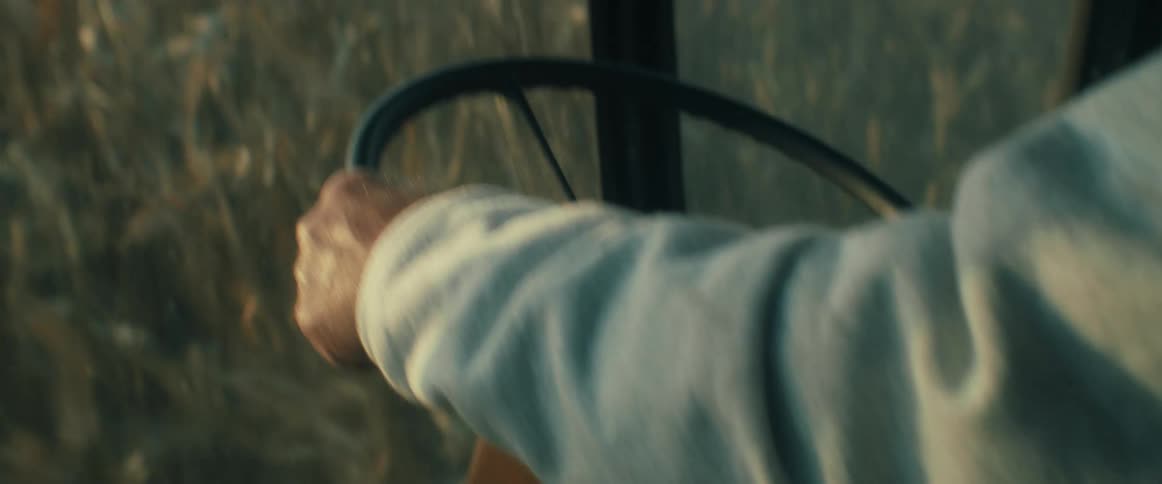 A close-up profile of a determined older farmer driving a combine harvester through a vast cornfield at golden hour