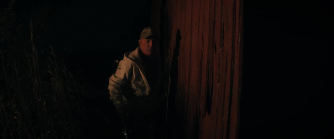 A man in a hoodie cautiously emerges from behind a red barn at night, illuminated by a warm glow