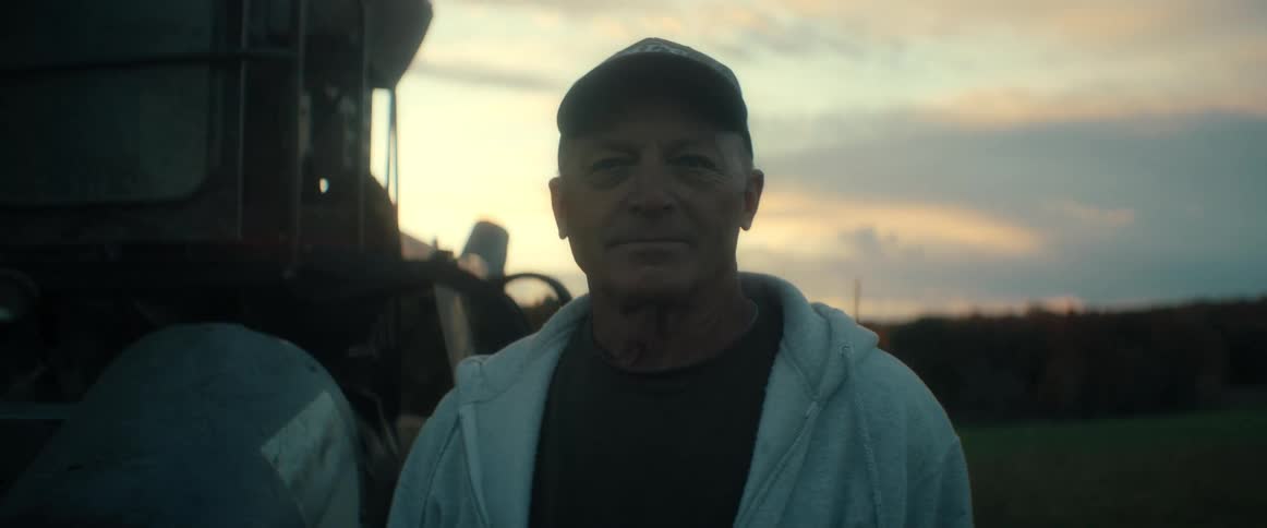A portrait of an older farmer standing proudly in his field next to his tractor at sunset