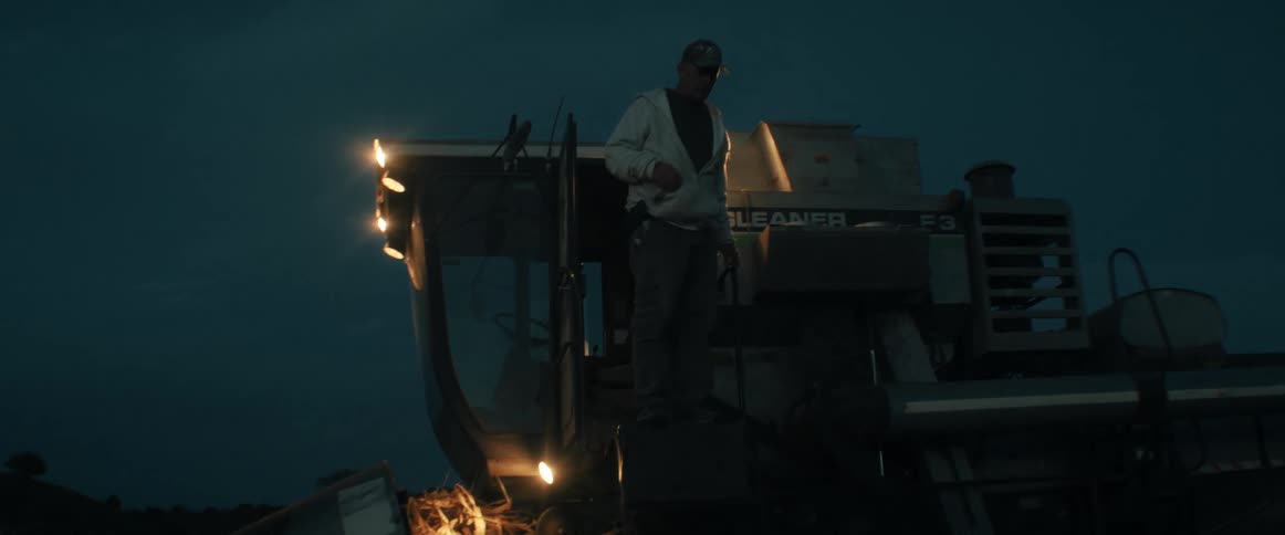 A farmer stands on his combine harvester at night before carefully climbing down into the harvested field