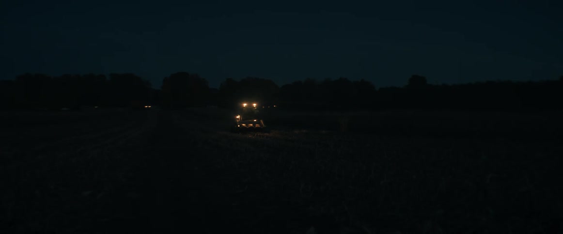 A tractor's headlights cut through the darkness as it harvests a crop in a rural field at night
