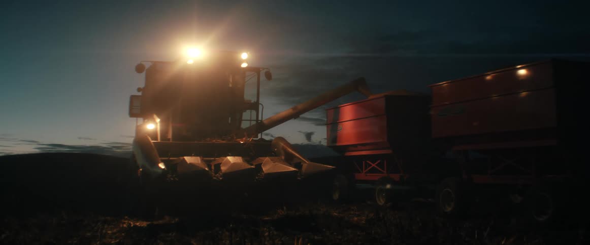 A powerful combine harvester with its lights on works late, unloading harvested grain from its auger into a red wagon at dusk