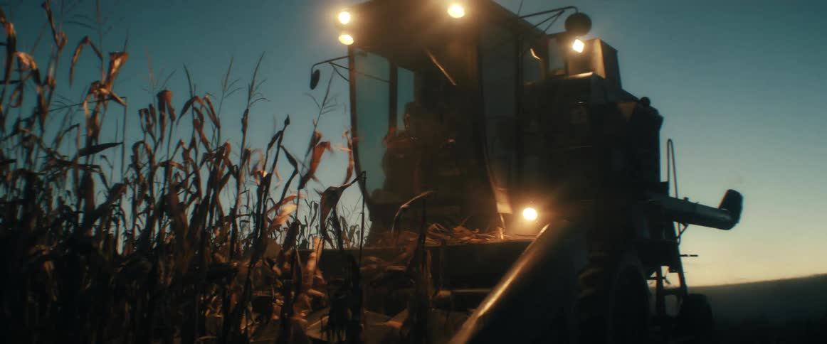 A combine harvester with bright lights on moves through a dry cornfield at dusk in slow motion