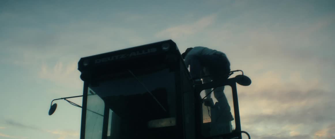 A farmer in a hoodie and baseball cap climbs onto his combine harvester and stands silhouetted against the sunset
