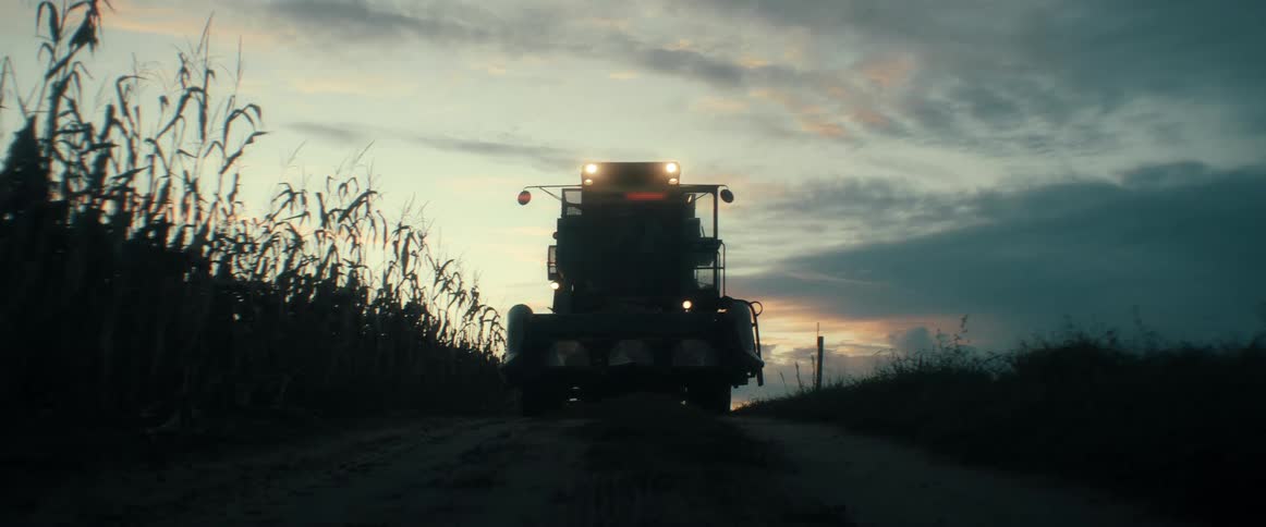 A massive combine harvester powers up its lights on a rural dirt road at dusk, preparing for the late-night corn harvest