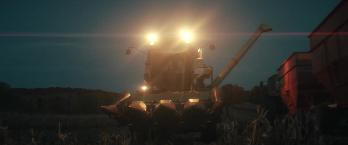 With bright headlights piercing the twilight, a combine harvester works late into the night, harvesting a cornfield and offloading grain