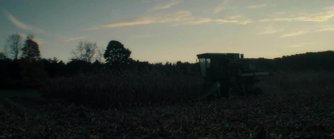 A combine harvester moves slowly through a rural cornfield, harvesting crops at sunset