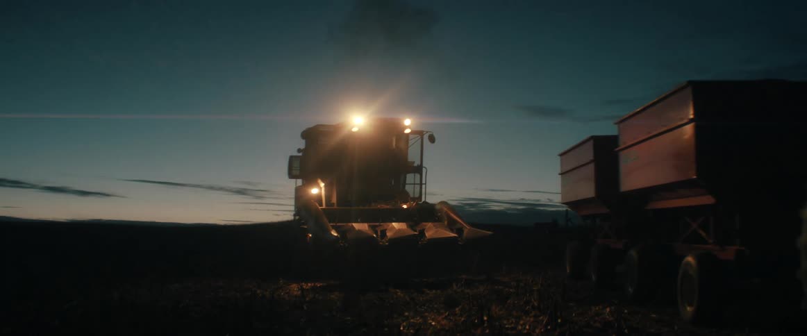 A combine harvester works late into the night, its bright headlights illuminating the field as it harvests crops