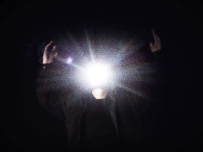 A grainy, vintage-style clip shows a surreal figure with a blinding, starburst light for a face raising their hands in the darkness