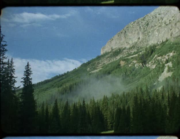 A vintage film shot captures a majestic mountain slope covered in a lush green forest with a light mist rising from the valley