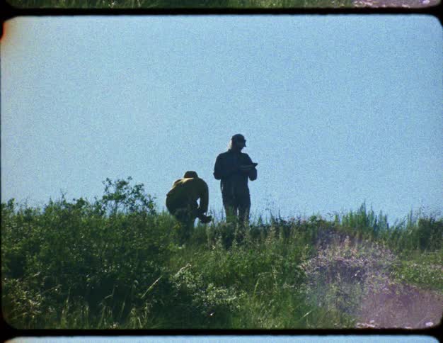 Archival footage from the 1970s shows two men, possibly geologists or surveyors, conducting fieldwork on a grassy hillside on a sunny day