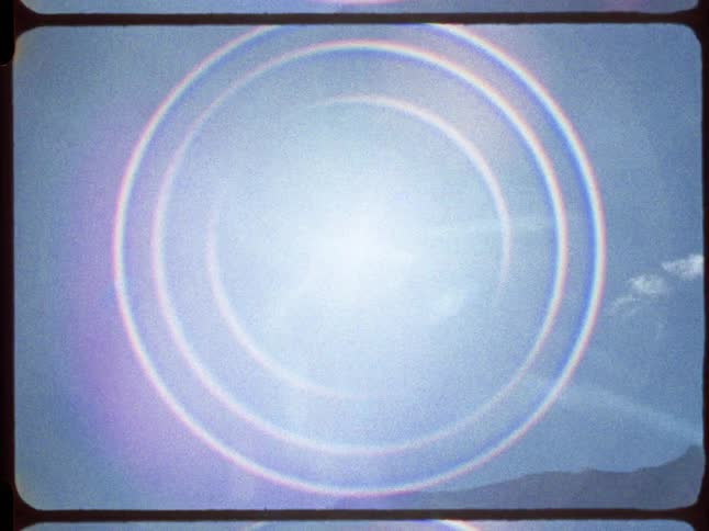 A vintage 8mm film captures a dreamy lens flare in the blue sky before tilting down to reveal a lush green mountain landscape