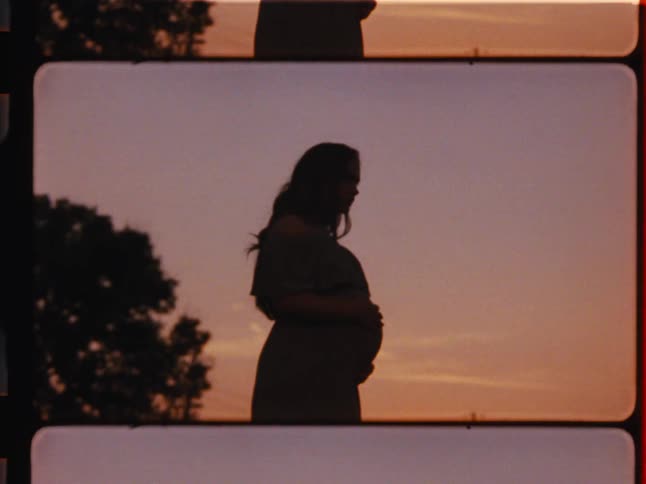 Vintage film shot of a silhouetted pregnant woman gently holding her belly against a warm sunset sky