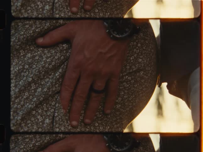 Vertical film-style close-up of an expecting father's hand on his wife's belly as he leans in to give it a loving kiss