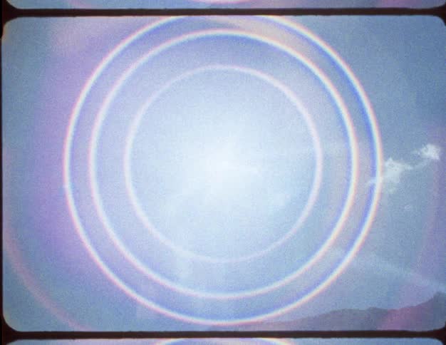Super 8 footage shows a psychedelic circular rainbow lens flare forming around the bright sun in the blue sky