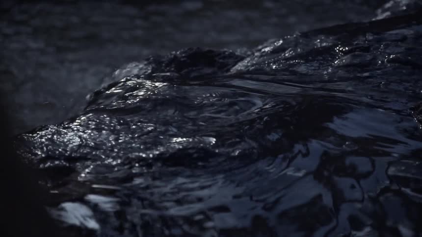 A moody close-up shot in slow motion captures dark, reflective water flowing gently over a submerged rock