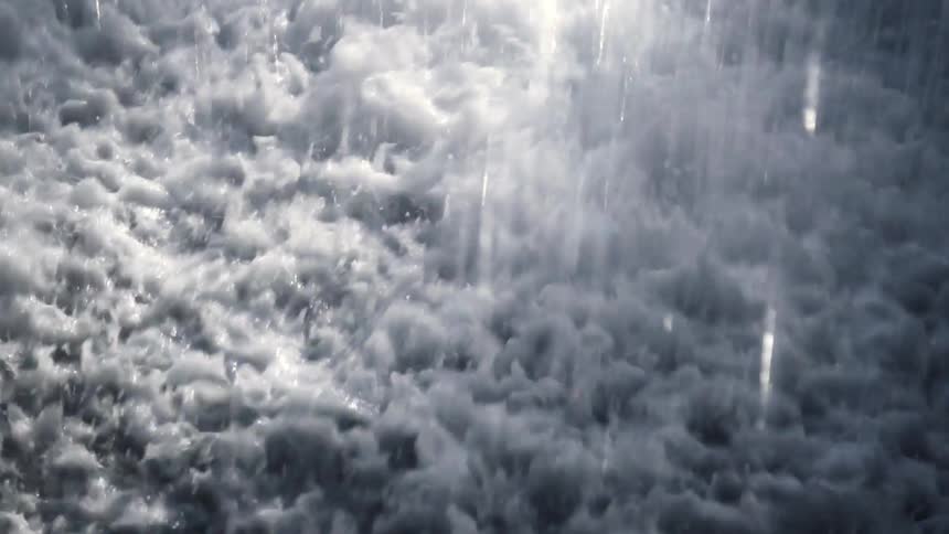 A dramatic slow-motion close-up captures the turbulent, foamy surface of water as it's pelted by falling streams