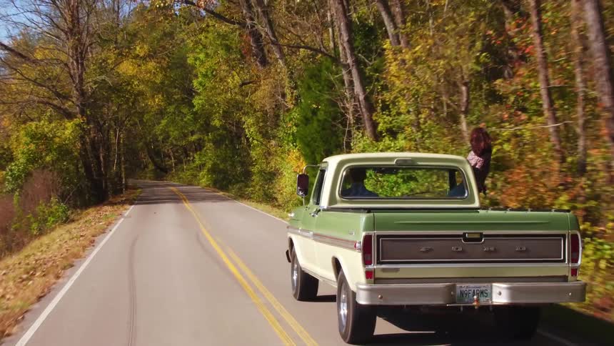 An old pickup truck drives through a forested area in the fall