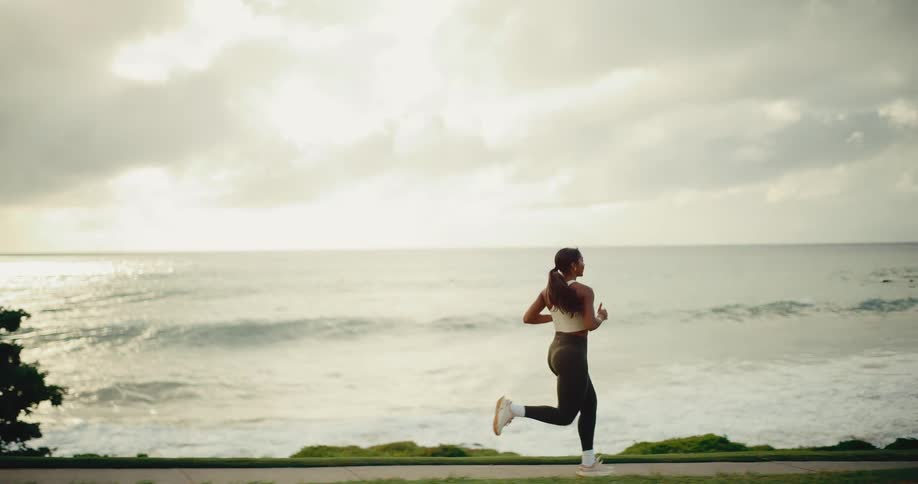 Side view of a woman in athletic wear jogging along a path by the ocean at dawn in slow motion