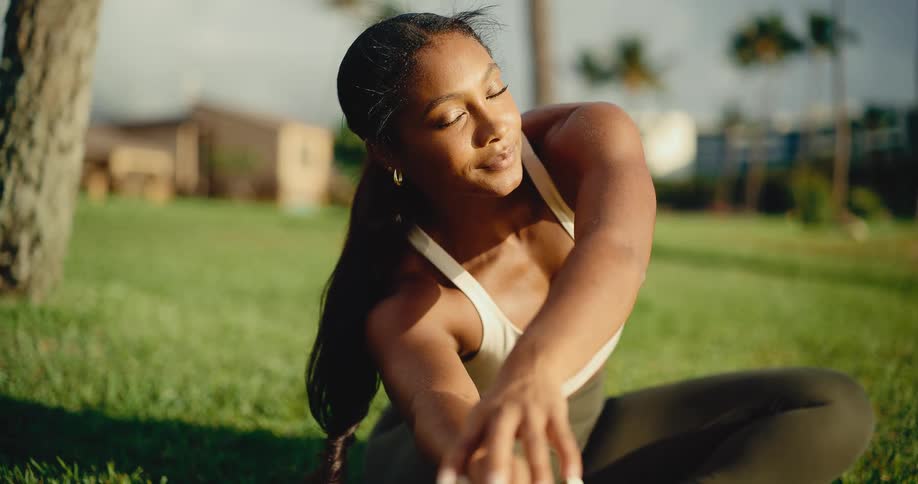 A serene athletic woman stretches deeply while sitting on the lush green grass of a park in the golden morning light