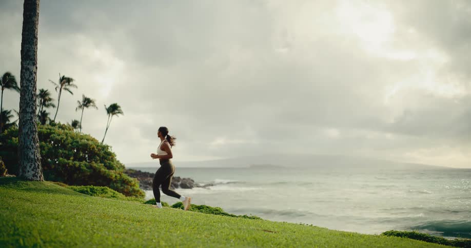 An athletic woman runs in slow motion along a grassy coastline with palm trees and the ocean in the background at sunrise