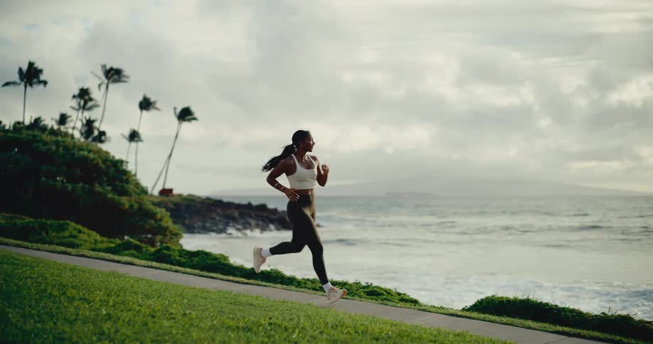An athletic woman runs in slow motion on a paved path next to the ocean with palm trees in the background