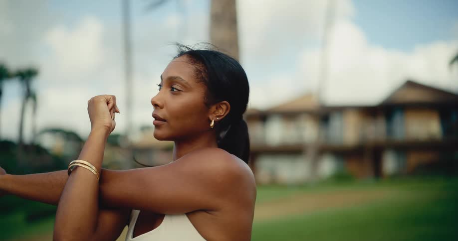 A serene woman stretches her arms in slow motion, warming up outdoors in a tropical park