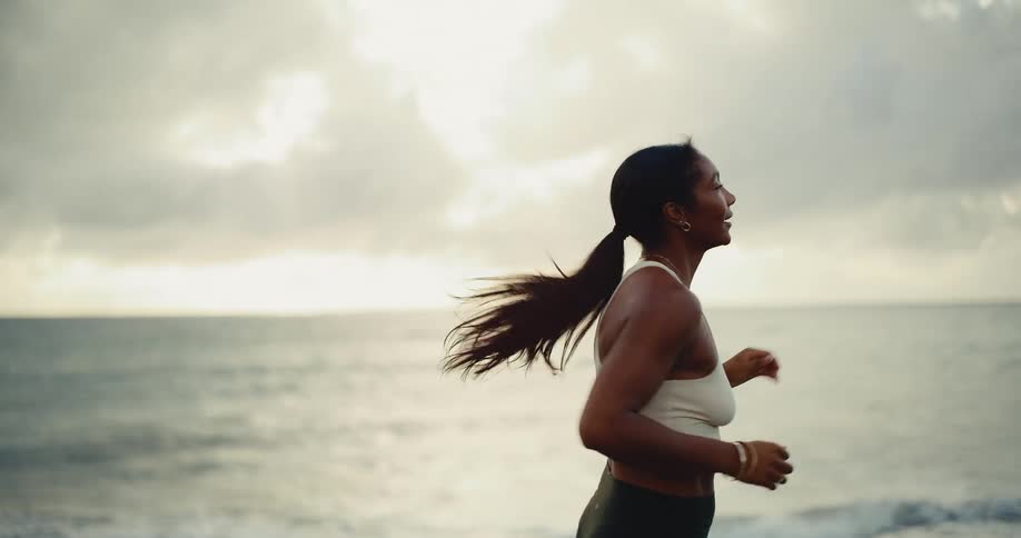 A fit athletic woman with a ponytail runs in slow motion with the ocean and a cloudy sunrise in the background