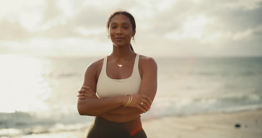 A beautiful and fit woman in athletic wear smiles confidently while standing on the beach at sunrise with the ocean in the background