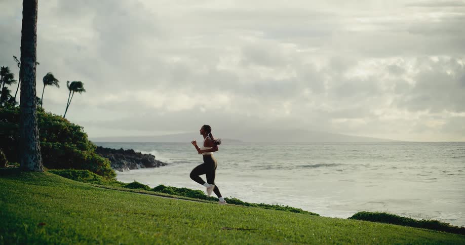 An athletic woman runs in slow motion on a grassy path along the tropical coastline at sunrise