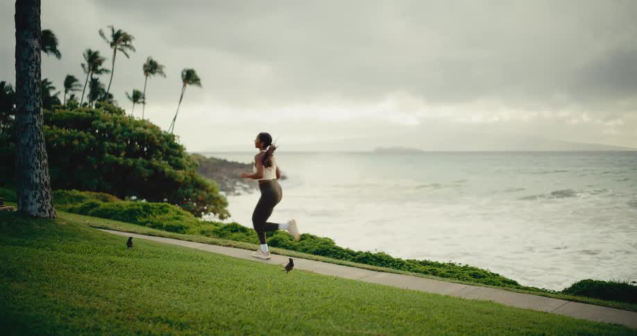 An athletic woman runs in slow motion along a beautiful coastal path with ocean waves crashing in the background