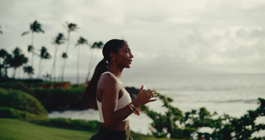 A beautiful Black woman jogs in slow motion along a tropical coastline at sunrise
