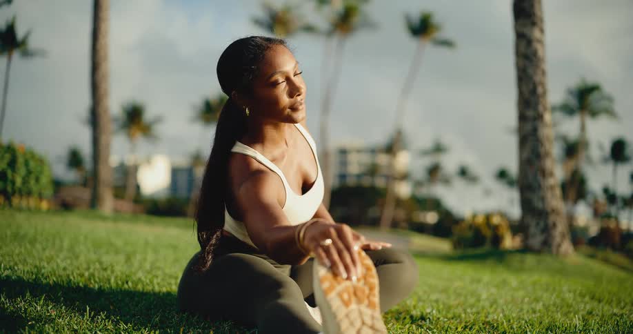 A beautiful, athletic woman in a white top stretches her legs while sitting in a grassy park surrounded by palm trees in slow motion