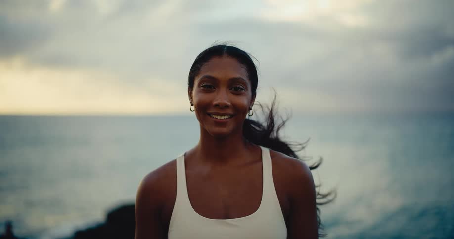 Slow motion portrait of a beautiful young woman smiling by the ocean with her hair blowing in the wind at dusk