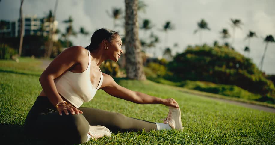 A fit Black woman performs a relaxing leg stretch in a beautiful park with the ocean and palm trees in the background during golden hour