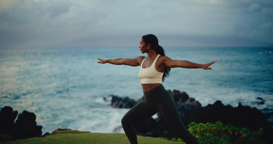 Woman gracefully practicing yoga on a grassy cliffside with a beautiful ocean view in slow motion