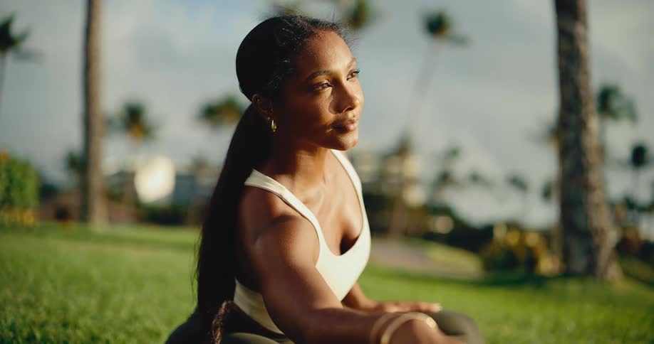 Close-up of a serene athletic woman practicing a yoga twist stretch on a lush green lawn surrounded by palm trees