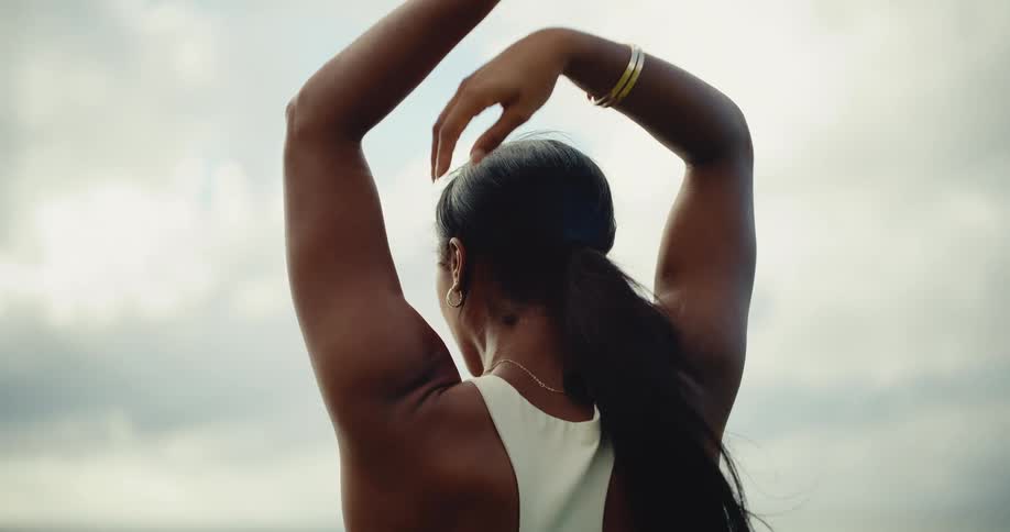 Close up from behind of a woman with long dark hair stretching her arms outdoors