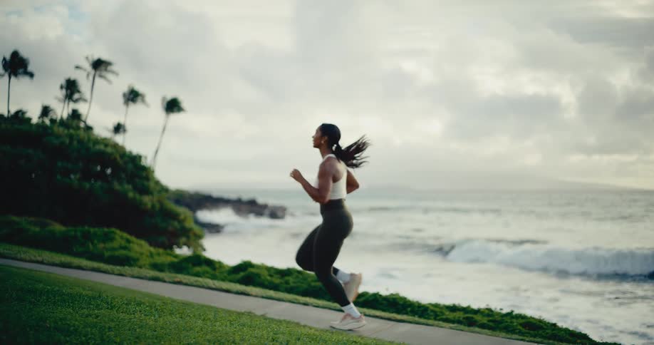 An athletic woman runs in slow motion along a path on a tropical coastline with ocean waves in the background