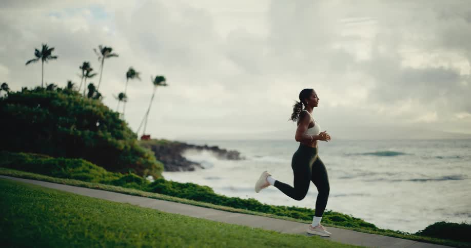 Slow motion shot of an athletic Black woman jogging on a beautiful path by the ocean at sunrise
