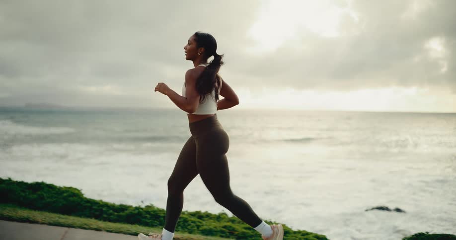 Slow motion shot of an athletic woman jogging along a beautiful coastal path with the ocean waves in the background at sunrise