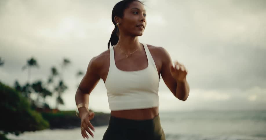 Slow motion shot of a beautiful athletic woman running in a park by the ocean with palm trees at dusk