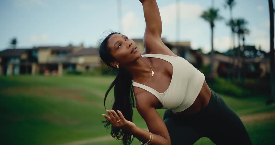 A graceful and serene woman flows through yoga poses outdoors on a lush green lawn in a tropical setting