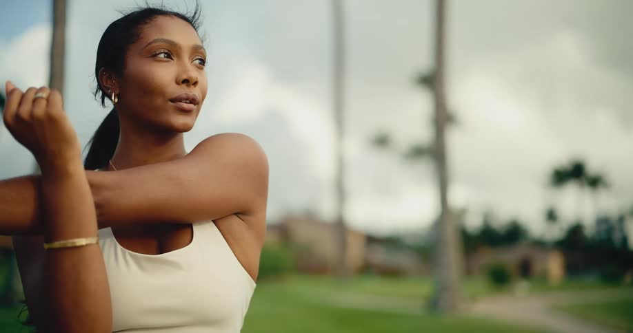 A beautiful Black woman in a white athletic top gracefully stretches her arms and shoulders in a tropical park, warming up for her workout