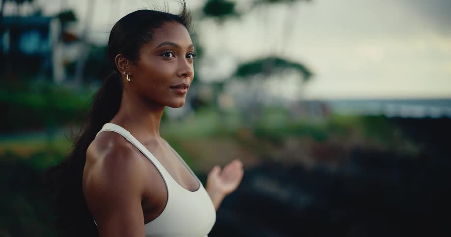 A beautiful, serene woman in athletic wear looks out at the ocean from a rocky shore at dusk before turning to look at the camera