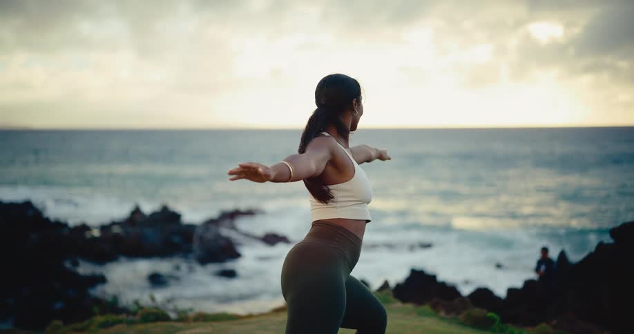 A woman gracefully practices yoga in slow motion on a grassy cliff overlooking the ocean at sunset
