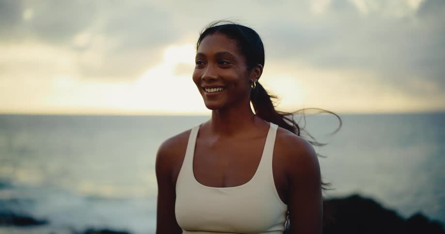 A beautiful athletic woman smiles peacefully by the ocean at sunset as the wind blows through her hair